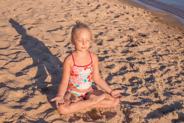 Een klein meisje in de lotuspositie op het zand mediteert 's avonds bij zonsondergang. Zomervakantie