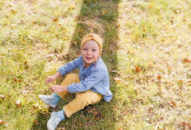 Een klein meisje in een blauw shirt en een gele broek zit op het gras in het park en glimlacht opzoeken