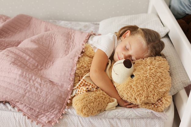 een klein meisje in een blond T-shirt slaapt thuis onder een roze deken en knuffelt een teddybeer