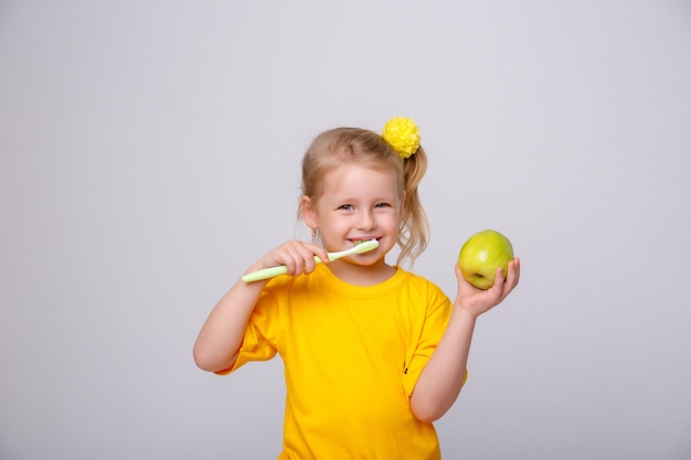 Een klein meisje in een geel T-shirt houdt een tandenborstel en een appel vast op een witte achtergrond