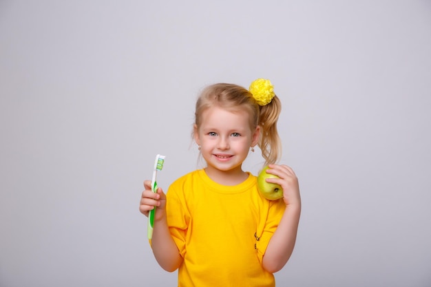 Een klein meisje in een geel T-shirt houdt een tandenborstel en een appel vast op een witte achtergrond