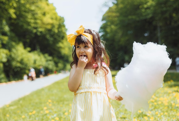 Een klein meisje in een gele jurk tijdens een wandeling in een pretpark eet suikerspin