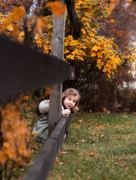 Een klein meisje in een geruite jurk kijkt uit vanaf een houten hek in een herfstpark.