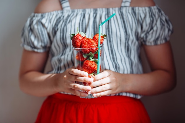 Een klein meisje in een gestreepte blouse en een rode rok houdt een glas met sappige aardbeien vast