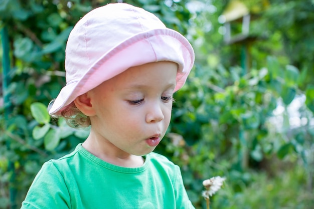 Foto een klein meisje in een groen t-shirt en een roze panama loopt in de zomer in het park en blaast de