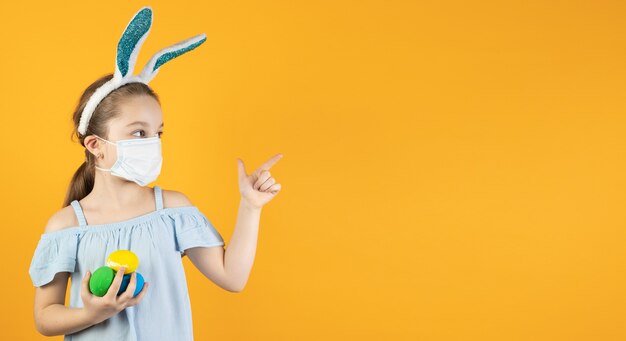 Een klein meisje in een medisch masker voor coronavirus op haar gezicht, op haar hoofd met konijnenoren, houdt paaseieren in haar handen, wijst met haar vinger naar een lege ruimte voor tekst