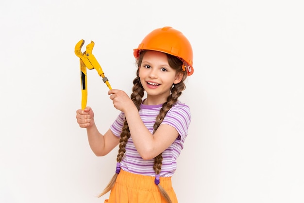 Een klein meisje in een oranje bouwhelm houdt een moersleutel vast op een witte geïsoleerde achtergrond Het concept van renovatie in de kinderkamer