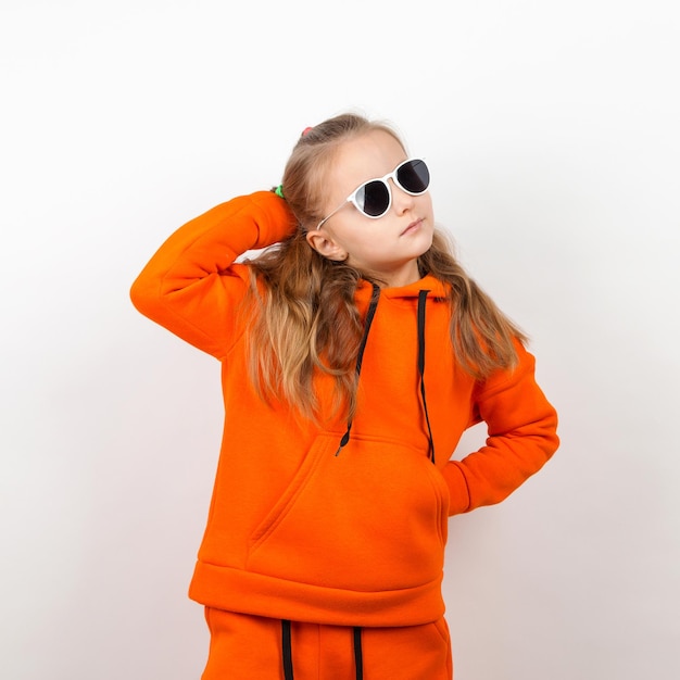 Een klein meisje in een oranje hoody en zonnebril emoties van vreugde en een zonnig dagportret op een witte achtergrond