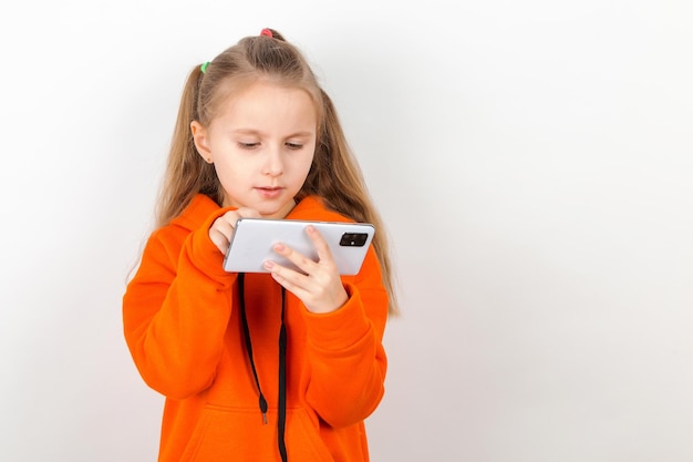 Een klein meisje in een oranje pak kijkt naar haar telefoon afhankelijkheid van een generatie kinderen op gadgets op een witte achtergrond