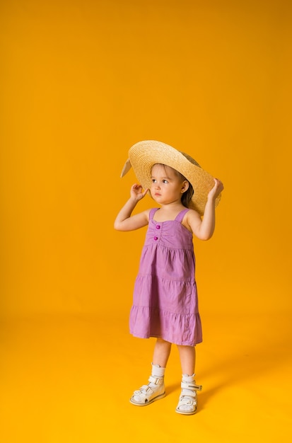 Een klein meisje in een paarse zomerjurk en een strooien hoed op een gele ondergrond met ruimte voor tekst