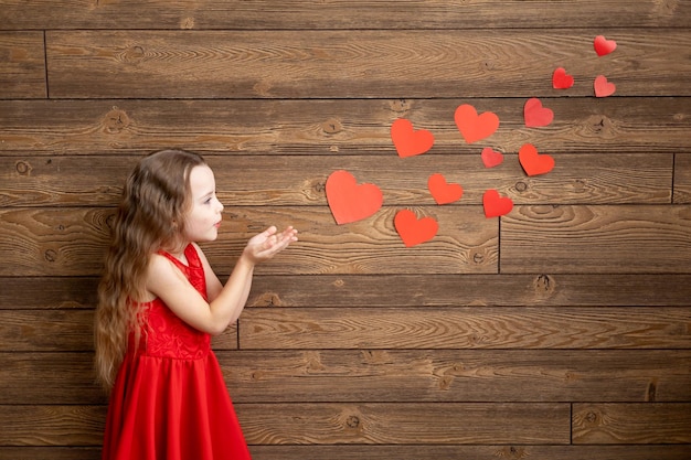 Een klein meisje in een rode jurk blaast harten uit haar handen op een donkerbruine houten achtergrond het concept van Valentijnsdag een lege ruimte voor tekst
