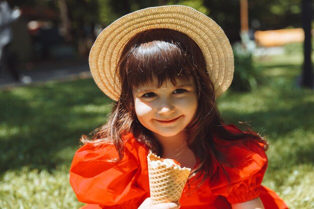 Een klein meisje in een rode jurk en een strohoed eet ijs in een kegel in het park