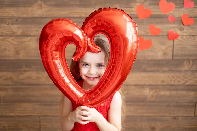 Een klein meisje in een rode jurk houdt een grote ballon in de vorm van een hart op een donkerbruine houten achtergrond met rode harten het concept van Valentijnsdag een plek voor tekst