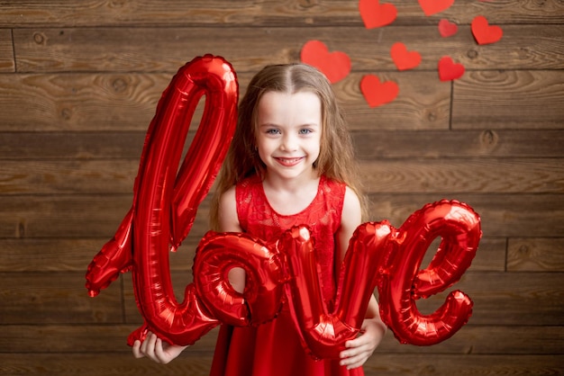 Een klein meisje in een rode jurk houdt in haar handen een grote ballon met de inscriptie Love op een donkerbruine houten achtergrond met rode harten het concept van Valentijnsdag