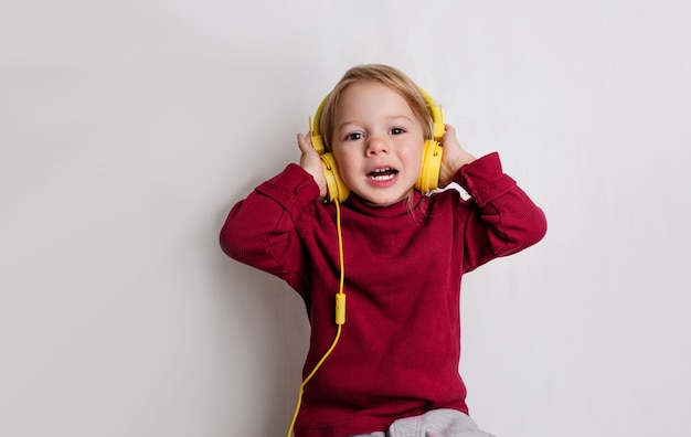 Een klein meisje in een rode trui luistert naar muziek met een koptelefoon en lacht op een witte achtergrond.