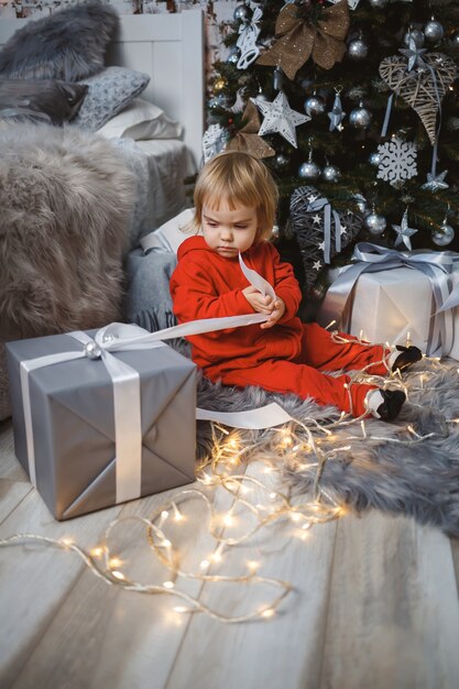 Een klein meisje in een rode warme trui zit onder een kerstboom met speelgoed en cadeaus. Gelukkige jeugd. Nieuwjaars vakantie sfeer