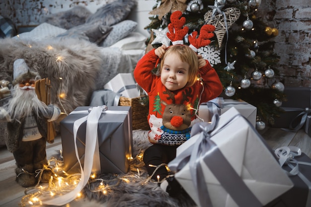 Een klein meisje in een rode warme trui zit onder een kerstboom met speelgoed en geschenken met hoorns op haar hoofd. Gelukkige jeugd. Nieuwjaars vakantie sfeer