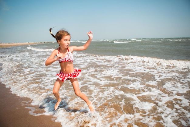 Een klein meisje in een rood zwempak speelt op het strand met een zeegolf die springt en plezier heeft Zwemmen reizen spelen met water