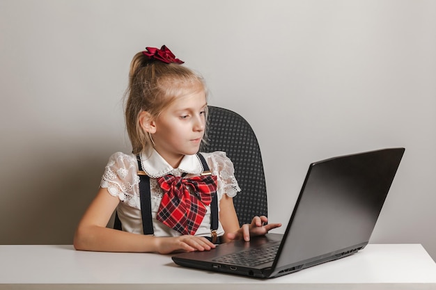 Een klein meisje in een schooluniform gebruikt een laptop aan een tafel op een witte achtergrond