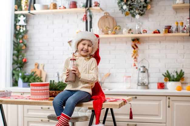 Een klein meisje in een trui en een kerstmuts, cacaomelk drinken zittend op de keukentafel.