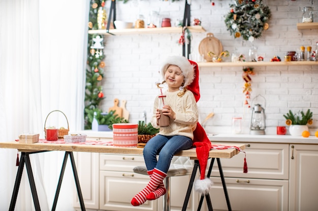 Een klein meisje in een trui en een kerstmuts, cacaomelk drinken zittend op de keukentafel.