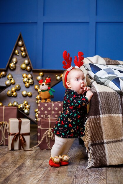 Een klein meisje in een warme trui zit onder een kerstboom met speelgoed en geschenken met hoorns op haar hoofd. Gelukkige jeugd. Nieuwjaars vakantie sfeer