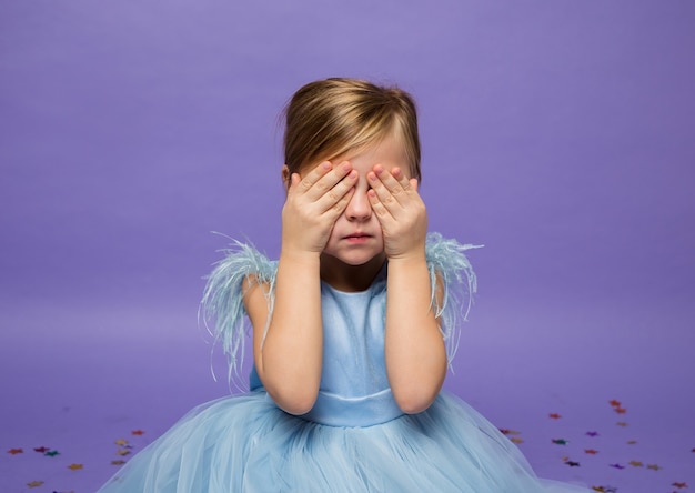 Een klein meisje in een weelderige blauwe jurk bedekte haar ogen met haar handen op paars