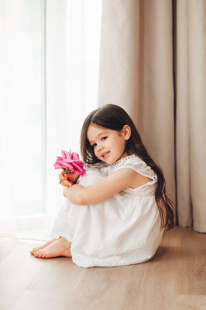 Een klein meisje in een witte jurk zit bij het raam en houdt een roos vast Een gelukkig kind bij het raam met een bloem