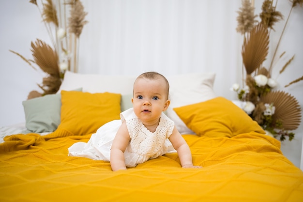 Een klein meisje in een witte katoenen jurk kruipt op een bed met een gele sprei