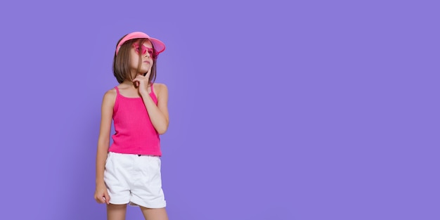 Een klein meisje in een witte korte broek, een roze T-shirt, een roze trendy bril en een zomerklep