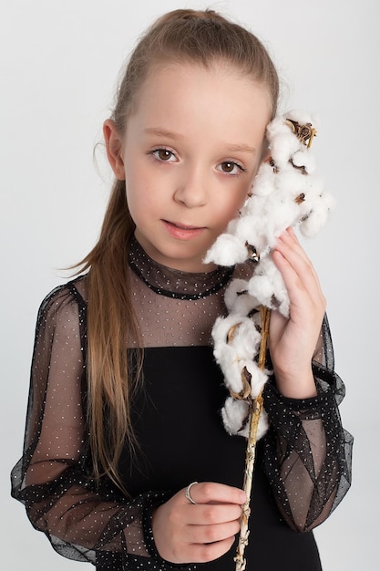 Een klein meisje in een zwarte jurk met een katoenen tak in haar handen staat in de studio op een witte achtergrond en glimlacht