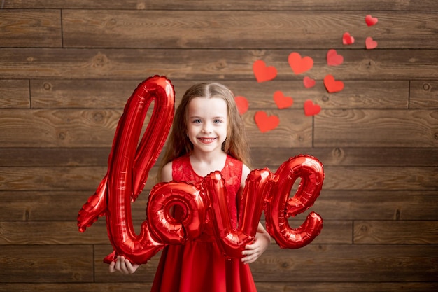 een klein meisje kind in rode pyjama met de inscriptie Liefde ligt op een donkerbruine houten achtergrond op de vloer en lacht met glazen in de vorm van harten het concept van Valentijnsdag