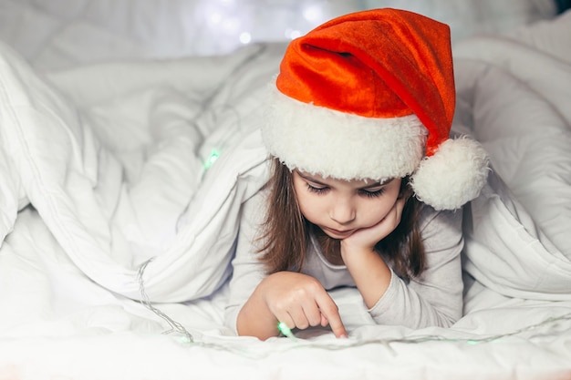 Een klein meisje ligt in bed in een kerstmuts en kijkt naar tekenfilms, studeert, speelt een spelletje aan de telefoon