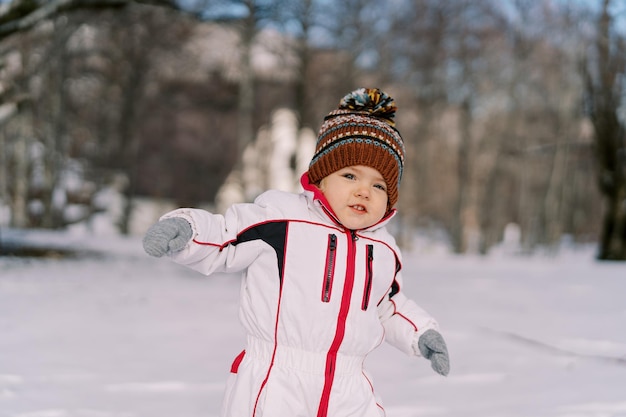 Een klein meisje loopt in de sneeuw in het bos en zwaait met haar handen.