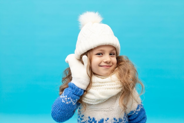 Een klein meisje luistert met haar hand naar haar oor op een blauwe achtergrond in een wintermuts en trui met wanten ruimte voor tekst