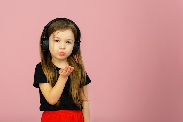 Een klein meisje luistert naar muziek in een grote koptelefoon op een roze pastelmuur en stuurt een kus naar de camera. Oprechte emoties