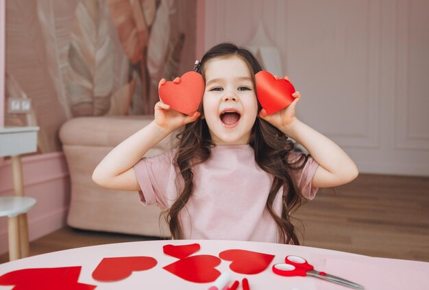 Een klein meisje maakt Valentijnsdagkaarten met gekleurd papier, een schaar en potlood zittend aan een tafel