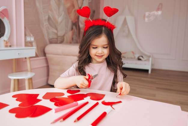 Een klein meisje maakt Valentijnsdagkaarten met gekleurd papier, een schaar en potlood zittend aan een tafel