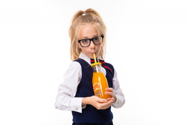 Een klein meisje met blond haar gevuld in een paardenstaart, grote blauwe ogen en een schattig gezicht in vierkante zwarte glazen drinkt sinaasappelsap in een glazen fles buis.
