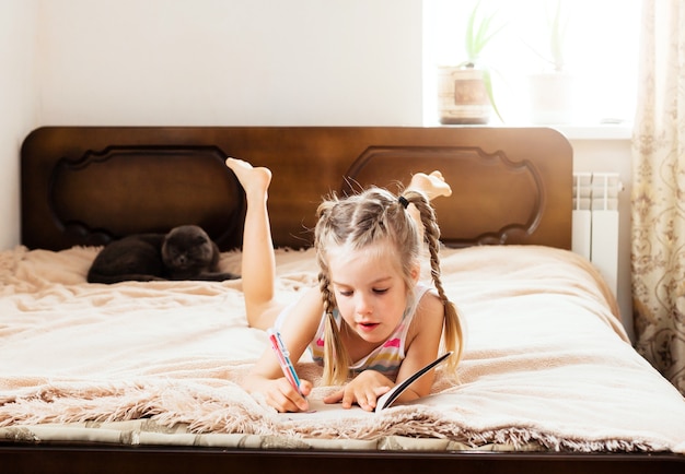 Een klein meisje met blond haar ligt thuis op een bed met een kat die een boek leest. Blijf thuis. Thuisonderwijs voor kinderen.