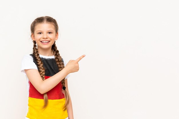 Een klein meisje met een Duitse vlag op een T-shirt met lang haar in staartjes wijst met haar wijsvinger zijwaarts naar uw advertentie op een witte geïsoleerde achtergrond Taalcursussen Kopieer de ruimte