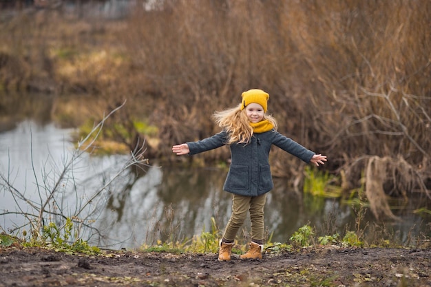 Een klein meisje met een gele hoed en sjaal loopt in de herfst langs de oever van een rivier