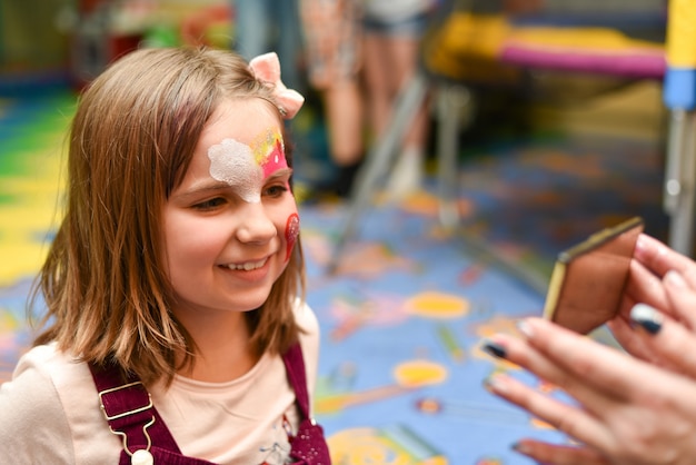 Een klein meisje met een geschilderd gezicht kijkt naar zichzelf in de spiegel op het feest