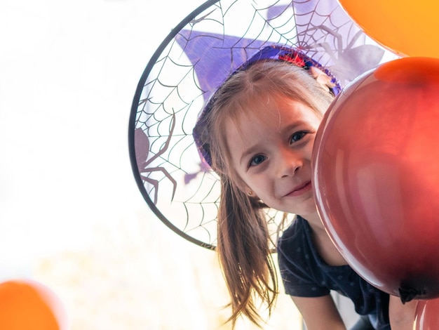 Een klein meisje met een heksenhoed gluurt achter een ballon en lacht vrolijk Halloween