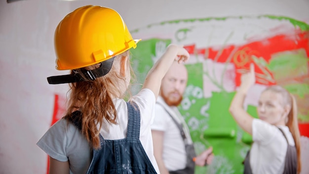 Een klein meisje met een helm wijst naar de muur, haar ouders staan naast de muur