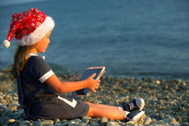 Een klein meisje met een kerstmanhoed zit aan de kust bij zonsondergang en speelt op de tablet