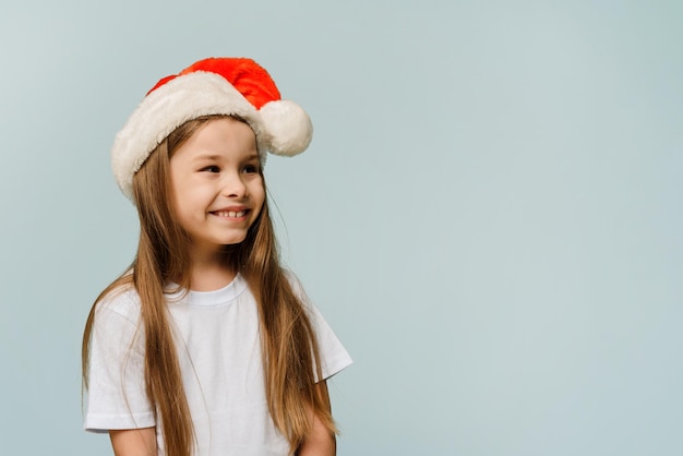 Een klein meisje met een kerstmuts kijkt zijwaarts op een blauwe achtergrond Kopieer ruimte Kerstmis en Nieuwjaar concept