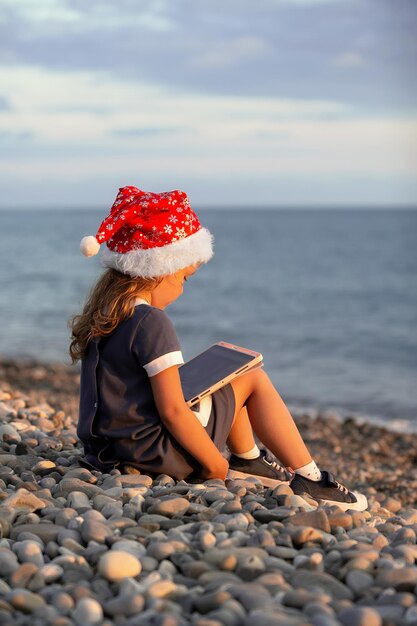 Een klein meisje met een kerstmuts zit aan de kust bij zonsondergang en kijkt naar de tekenfilms op tablets