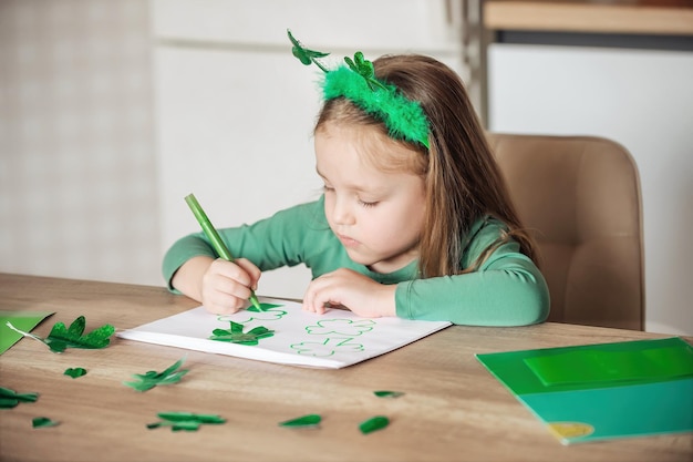 Een klein meisje met een klaverhoofdband tekent en snijdt groene klavers voor St. Patrick's Day