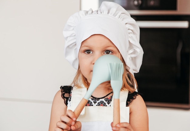 Een klein meisje met een koksmuts speelt met keukenapparatuur in de keuken